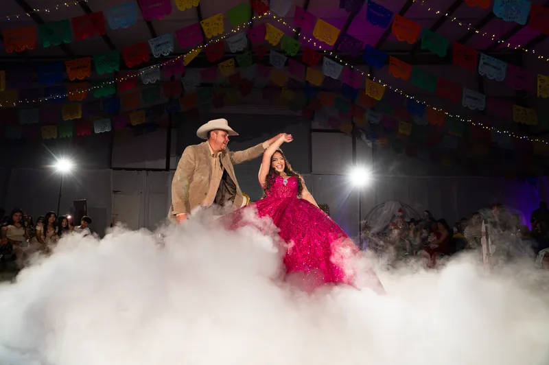 Father and daughter share a Dance on the Clouds moment on a fog-covered dance floor under papel picado banners at a DJ Oso quinceañera